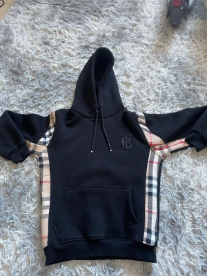 Burrbery Hoodie  - Snygg svart hoodie från Burberry med klassiska beige, röda och svarta rutiga paneler längs sidorna och ärmarna. Stor huva med dragsko och stor magficka framtill. Broderad TB-logga på bröstet. Perfekt för dig som vill ha en exklusiv och trendig look. Den är storlek M men känns som S.
