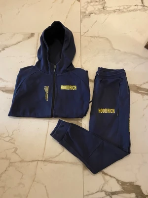 Marinblå Hoodrich tracksuit set-M - Snyggt marinblått tracksuit set från Hoodrich med gul logga på både hoodie och byxor. Hoodien har dragkedja och huva, byxorna har resår i midjan och smal passform. Perfekt för en avslappnad streetwear-look.
