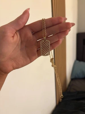 Guldigt halsband med rektangulär berlock - Snyggt halsband i guldfärg med en rektangulär berlock dekorerad med diagonala rader av små glittrande stenar. Kedjan är tunn och elegant, perfekt för att ge din outfit lite extra bling. Ingen synlig märkning på halsbandet.