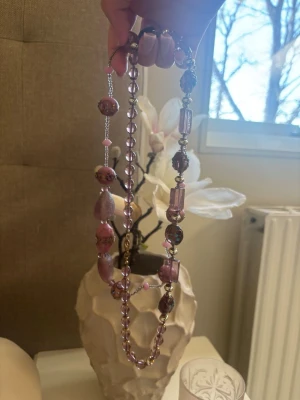Långt halsband med lila pärlor - Unikt långt halsband med olika former av lila och rosa pärlor i både runda, ovala och cylinderformade varianter. Halsbandet har en mix av transparenta och matta pärlor som ger en cool och lekfull look. Perfekt för att addera färg till din outfit.