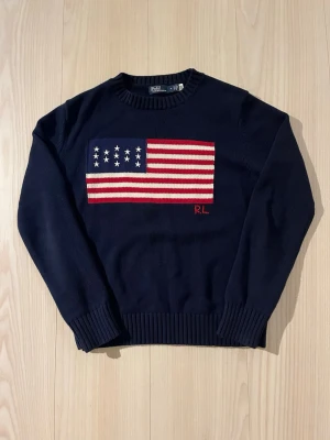 Mörkblå stickad tröja Polo Ralph Lauren - Mörkblå stickad tröja från Polo Ralph Lauren med amerikansk flagga i rött, vitt och blått på bröstet. Rund halsringning och ribbade muddar vid ärmslut och nederkant. RL-broderi i rött under flaggan. Klassisk och tidlös design.