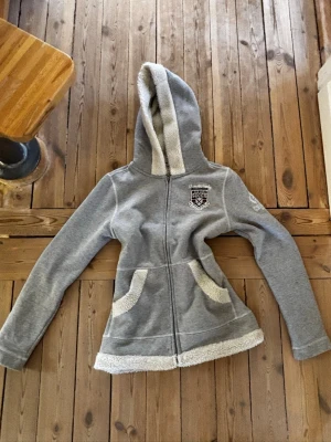 Grå hoodie med teddyfoder och brodyr - Mysig grå hoodie från Snoot med vitt teddyfoder i huva, fickor och nederkant. Dragkedja framtill, broderad logga på bröstet och snygg textdetalj på ryggen. Perfekt för chill dagar och streetwear-vibe. Har sytt in den själv, se bilder