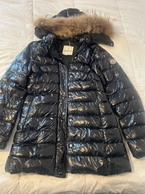 Svart glansig dunjacka från Moncler - Snygg svart dunjacka från Moncler med glansig finish och avtagbar huva med fuskpäls. Jackan har en längre modell, dragkedja och tryckknappar framtill samt Moncler-logga på ärmen. Perfekt för kalla vinterdagar och riktigt varm.