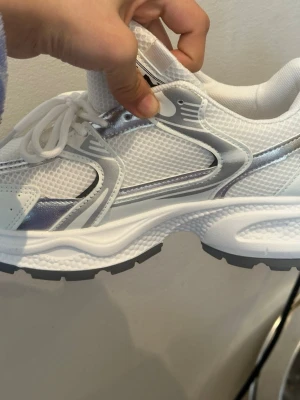 Vita chunky sneakers med silverdetaljer - Snygga vita sneakers med chunky sula och silvriga detaljer. Ovandelen är i mesh och syntetmaterial för en sportig look. Skorna har vita snören och grå yttersula. Perfekta för dig som gillar trendiga och bekväma sneakers.