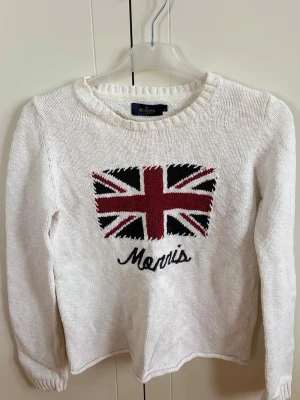 Vit stickad tröja Morris Lady - Vit stickad tröja från Morris Lady med Union Jack-flagga i rött, svart och vitt på bröstet samt Morris-broderi under. Tröjan har rund halsringning och långa ärmar. Perfekt för dig som gillar klassisk brittisk stil och vill ha något bekvämt och snyggt.