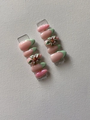 Press-on naglar med 3D-blommor - Säljer ett set press-on naglar i ljusrosa med mintgröna tippar och detaljerade 3D-blommor. Naglarna har en oval form och blommorna har gulddetaljer i mitten för extra wow-effekt. Perfekt för dig som vill ha en söt och unik nagelstil.