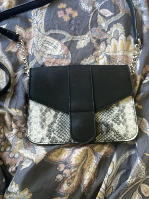 Svart axelväska med ormmönster - Snygg axelväska i svart konstläder med detaljer i ormmönstrat material. Väskan har silverfärgade kedjor på axelremmen och en unik design på locket. Perfekt för dig som vill ha en trendig accessoar med lite edge.
