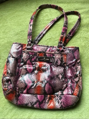 Handväska från Nine West - Säljer en unik handväska från Nine West med färgstarkt ormmönster i lila, rosa och svart. Väskan har dubbla axelband, flera innerfack och dragkedja. Materialet är konstläder med glansig finish och väskan har en metallbricka framtill. Tag 🏷️ på