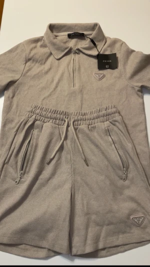 Prada set beige polo shorts tröja - Helt ny aldrig använd. Snygg beige pikétröja från Prada med diskret rutmönster och ikonisk metallbadge på bröstet. Tröjan har krage, korta ärmar och en stilren half zip framtill. Perfekt för dig som gillar clean och lyxig design. Kommer med shorts med zip fickor.