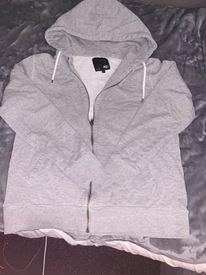Ljusgrå hoodie med dragkedja 157 - Ljusgrå hoodie från 157 i storlek XL. Klassisk modell med dragkedja framtill, huva med vita snören och mjukt, borstat innerfoder. Perfekt för chill dagar eller när du vill ha en enkel och clean look. Materialet är mjukt och bekvämt.