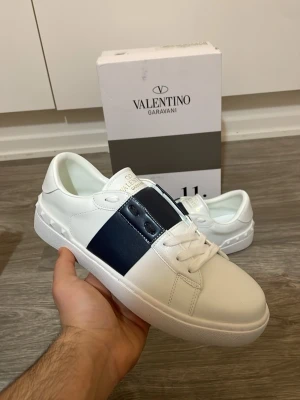 Valentino open skor - Snygga vita sneakers från Valentino Garavani med bred marinblå detalj över snörningen. Skorna har rund tå, platt sula och är tillverkade i skinn. Perfekta för dig som vill ha en clean och exklusiv look.