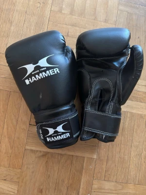 Svarta boxningshandskar från Hammer - Säljer ett par svarta boxningshandskar från Hammer med vit logga och kardborreband runt handleden. Handskarna är tillverkade i slitstarkt syntetmaterial och har vit kontrastsöm. Perfekta för träning inom boxning eller kampsport.