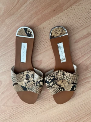 Ormmönstrade sandaler från Zara Basic - Snygga slip-in sandaler från Zara Basic med trendigt ormmönster i beige, brunt och svart. Sandalerna har breda korslagda remmar i konstläder och öppen tå. Perfekta för en cool och avslappnad look till sommaren.