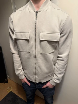 Beige jacka med fickor. - Snygg beige bomberjacka med två stora bröstfickor och dragkedja framtill. Jackan har en clean look, ribbade muddar och lång ärm. Perfekt för dig som gillar minimalistisk och clean stil. Nypris: 500. Använd 5 gånger. Storlek: S.