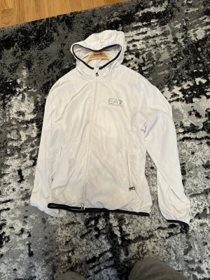 Vit vindjacka från EA7 Emporio Armani - Snygg vit vindjacka från EA7 Emporio Armani med huva och dragkedja framtill. Jackan har två fickor med dragkedja och meshfoder på insidan för extra komfort. Perfekt för dig som vill ha en stilren och sportig look.