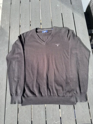 Svart stickad tröja från GANT - Klassisk svart stickad tröja från GANT med V-ringning och diskret broderad logga på bröstet. Tröjan har ribbade muddar vid ärmslut och nederkant, vilket ger en snygg passform. Perfekt att matcha med skjorta eller t-shirt.