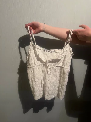 Vit broderad somrig topp - Söt vit topp med tunna axelband och broderade blommor. Modellen har volangkant upptill och en liten knytning framtill. Perfekt till sommaren. Luftig och lätt, passar till både jeans och kjol.