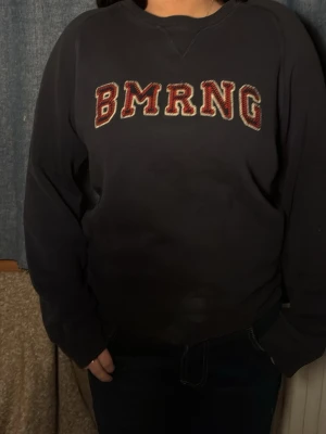 Svart sweatshirt med BMRNG-tryck - Cool svart sweatshirt med stora bokstäver 'BMRNG' i rött och vitt framtill. Tröjan har rund halsringning och långa ärmar. Perfekt för en avslappnad streetwear-look.