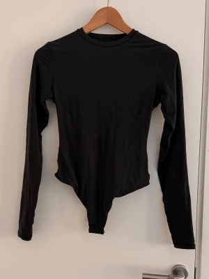 SKIMS Fits Everybody Long Sleeve Crew Neck Bodysuit - Onyx (Stl S) - SKIMS Fits Everybody långärmad bodysuit med rund hals i färgen "Onyx" (klassisk svart). • Storlek: Small (S) • Skick: Mycket gott skick, sparsamt använd. • Material: Extremt mjukt, stretchigt och följsamt tyg (76% polyamid, 24% elastan). • Detaljer: Rund halslinning, thong-skärning baktill för att undvika synliga kanter under kläderna, samt knäppning i grenen. Detta är den virala bodyn som är känd för sin fantastiska passform som formar sig efter kroppen. 