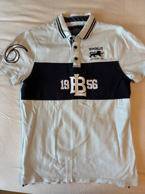Ljusblå piké från Bondelid - Snygg pikétröja från Bondelid i vitt och marinblått med broderad logga och texten 1956 på bröstet. Tröjan har krage med knappar och korta ärmar samt dekorativt tryck på ena ärmen. Perfekt för en sportig och avslappnad stil.