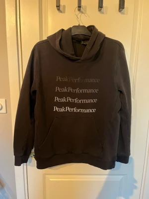 Svart hoodie Peak Performance L - Snygg svart hoodie från Peak Performance med stor huva och känguruficka. Framsidan har flera Peak Performance-loggor tryckta i vitt och grått. Tillverkad i mjukt bomullsmaterial som är skönt mot huden. Perfekt för en avslappnad och sportig stil.