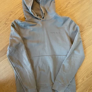 Grå hoodie från J.Lindeberg - Snygg grå hoodie från J.Lindeberg med broderad logga på bröstet. Klassisk modell med huva, känguruficka och långa ärmar. Perfekt för en chill och stilren look. Tillverkad i mjuk bomull för extra komfort. 