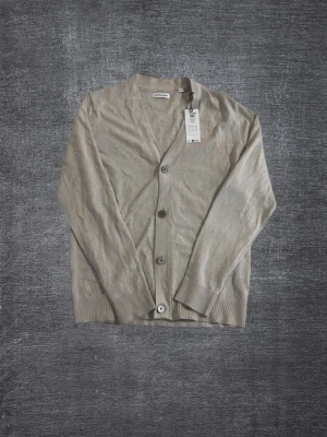 Beige cardigan från Jack & Jones NYSKICK - Stilren beige cardigan från Jack and Jones med v-ringning och knäppning framtill. Tillverkad i tunt, mjukt material med ribbade muddar vid ärmslut och nederkant. Perfekt för lager på lager och enkel att matcha med olika outfits.