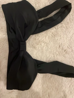 Svart bandeau bikini med breda band - Stilren svart bandeau-bikini med breda axelband och twistad detalj framtill. Bh:n är utan bygel och har en slät, mjuk yta i stretchigt material som ger en bekväm passform. Perfekt för dig som gillar minimalistisk och trendig design. 
