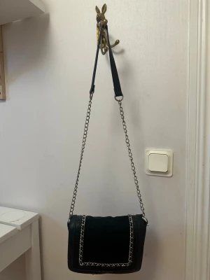 Svart axelväska med kedjedetaljer - Snygg svart axelväska med kedjerem och detaljer i silver. Väskan har en mix av mockaimitation och krokopräglat konstläder, vilket ger en cool kontrast. Perfekt storlek för det viktigaste och stängs med lock. Stilren och trendig design.