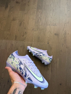 Nike Mercurial ljuslila fotbollsskor - Säljer ett par Nike Mercurial fotbollsskor i ljuslila med neon- och mörkblå detaljer. Skorna har en modern, strömlinjeformad design med mönstrad ovandel och en sula i genomskinlig lila och neon. Perfekta för dig som vill sticka ut på planen!