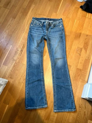 Blå bootcut jeans från Miss Me - Snygga blå bootcut jeans från Miss Me med klassiska fem fickor och dekorativa knappar. Jeansen har en lätt tvättad look och markerade sömmar som ger en cool vintagekänsla. Perfekta för dig som gillar en avslappnad men trendig stil.
