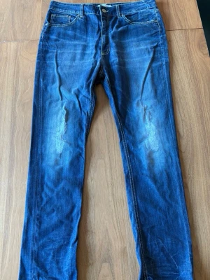Blå jeans från Dondup - Säljer ett par blå jeans från Dondup med snyggt slitna detaljer och klassisk femficksmodell. Jeansen har raka ben och kontrastsömmar som ger en cool look. Tillverkade i mjuk denim som sitter skönt hela dagen.