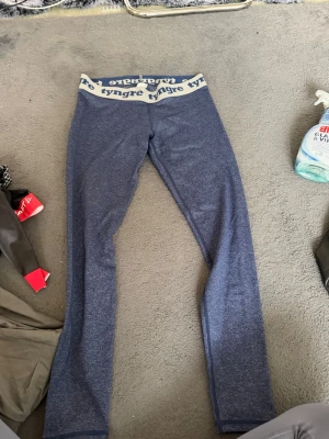 Blå leggings från Tyngre - Säljer ett par blå leggings från Tyngre med bred resår i midjan där loggan står tryckt i vitt. De har en tight passform och är tillverkade i ett stretchigt material som passar perfekt till träning eller chill. Snygg och enkel design som är lätt att matcha.