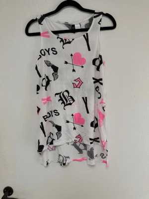 Vit linne med rosa hjärtan och tryck - Säljer ett vitt linne med svarta och rosa tryck, bland annat hjärtan med pilar, texten 'BOYS' och olika grafiska detaljer. Toppen är ärmlös och har en lös passform, perfekt för en avslappnad stil. Materialet känns lätt och luftigt. ( tänk på jag har hund i hemmet) 
