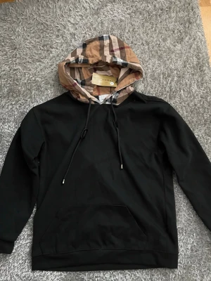 Svart Burberry hoodie - Snygg svart hoodie från Burberry med klassisk beige, röd och svart rutig huva. Hoodien har dragsko i huvan och en stor magficka framtill. Perfekt för dig som vill ha en stilren men ändå unik look. Materialet känns mjukt och bekvämt. Storlek L fast liten i storlek så passar M bättre. Helt ny och oanvänd så pris kan diskuteras vid snabb affär!