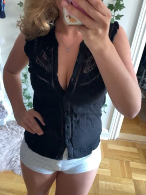 Svart ärmlös blus med spetsdetaljer - Snygg svart blus med spetsdetaljer upptill och knäppning framtill. Blusen är ärmlös och har en lätt v-ringad halsringning. Perfekt att styla till både jeans och kjol för en trendig look.