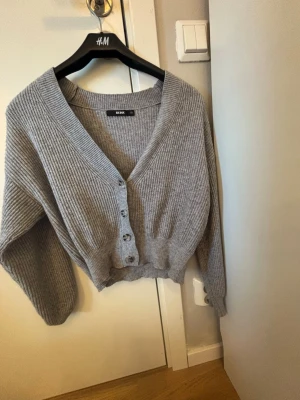 Grå ribbad kofta från Bik Bok - Mysig grå kofta från Bik Bok i ribbstickat material. Koftan har djup V-ringning, knappar framtill och en croppad passform. Perfekt att slänga över en topp för en avslappnad look.