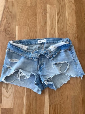 Ljusa denimshorts från Zara - Säljer ett par ljusblå denimshorts från Zara med råa kanter och slitna detaljer. Shortsen har fem fickor, bälteshällor och knappgylf. Perfekta för sommaren och ger en avslappnad vibe.