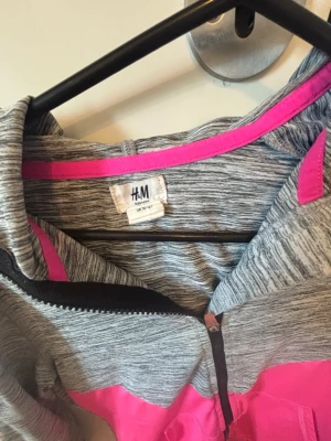 Gråmelerad hoodie från H&M - Sportig gråmelerad hoodie från H&M med dragkedja framtill och rosa detaljer. Långärmad modell med texten 'AWESOME' längs båda ärmarna. Tillverkad i mjukt och stretchigt material, perfekt för träning eller chill. Snygg huva och kontrastfärgade inslag.