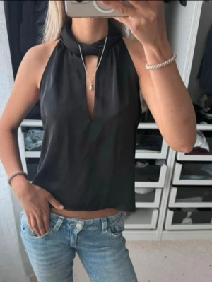 Svart halterneck festtopp Zara  - Snygg svart halterneck festtopp med knyt runt halsen och djup v-ringning framtill från Zara. Toppen är ärmlös och har en lös passform som ger en avslappnad men trendig look. Perfekt att matcha med jeans eller kjol för en cool stil. Bra skick!