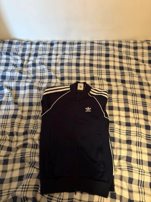 Marinblå Adidas ziptröja - Snygg marinblå ziptröja från Adidas med vita ränder på axlarna och vita detaljer längs ärmarna. Klassisk Adidas-logga på bröstet och dragkedja framtill. Perfekt för en sportig och avslappnad stil. Hoodie är xs 