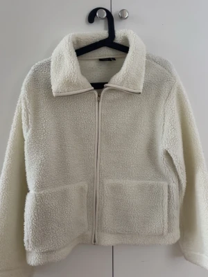 Vit teddyjacka med dragkedja - Mysig vit teddyjacka med hög krage och dragkedja framtill. Jackan har två stora fickor fram och är gjord i fluffigt, mjukt teddy-material. Perfekt för kalla dagar när du vill vara både varm och stilren.
