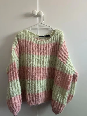 Pastellrandig stickad tröja - Mysig stickad tröja med breda pastellrosa och ljusgröna ränder. Tröjan har en lös passform, rund halsringning och långa ärmar. Materialet är fluffigt och ger en soft känsla, perfekt för att hålla värmen under kyliga dagar.