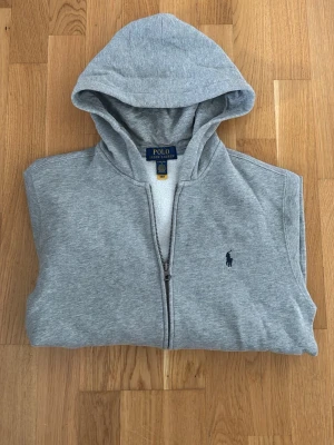 Grå hoodie från Polo Ralph Lauren - Snygg grå hoodie från Polo Ralph Lauren med dragkedja framtill och klassisk blå logga broderad på bröstet. Tillverkad i mjukt material och har en stor huva. Perfekt för en avslappnad och stilren look.