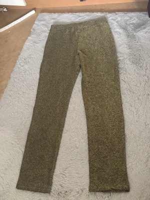 Glittriga guldleggings - Säljer ett par glittriga leggings i guld med rak passform och resår i midjan. Materialet är stretchigt och skimrar snyggt i ljuset, perfekt för dig som vill sticka ut lite extra. Inga fickor eller synliga detaljer, bara ren glitterkänsla. Aldrig använd, utan prislapp Indiska.