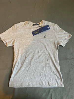 Grå t-shirt från Polo Ralph Lauren - Snygg grå t-shirt från Polo Ralph Lauren med klassisk passform och liten broderad logga på bröstet. Tillverkad i mjuk bomull som känns skön mot huden. Perfekt basplagg till alla outfits.