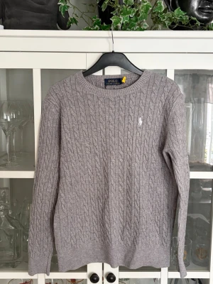 Grå kabelstickad tröja Polo Ralph Lauren - Snygg grå kabelstickad tröja från Polo Ralph Lauren med rund halsringning och den klassiska broderade loggan på bröstet. Tröjan har långa ärmar och ribbade muddar vid ärmslut och nederkant. Perfekt för lager på lager och stilrena outfits.