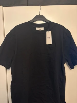 Svart t-shirt från Ami  - Klassisk svart t-shirt från Ami Paris med rund halsringning och diskret bröstficka. Tillverkad i mjuk bomull för en bekväm känsla och enkel stil. Perfekt basplagg för alla tillfällen och lätt att matcha med andra plagg.