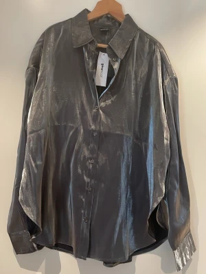 Silvrig oversized skjorta från Gina Tricot strl S - Säljer en glansig silvrig skjorta från Gina med oversized passform. Skjortan har klassisk krage, långa ärmar och knäppning framtill. Materialet är syntet och ger ett coolt metallic-utseende. Perfekt för dig som vill sticka ut med en unik look.