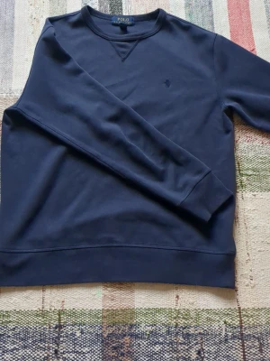Mörkblå sweatshirt från Polo Ralph Lauren - Klassisk mörkblå sweatshirt från Polo Ralph Lauren med rund halsringning och diskret broderad logga på bröstet. Tröjan har ribbade muddar vid ärmslut och nederkant, perfekt för en avslappnad och stilren look.